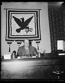 Black and white photo with man sitting at desk, Blue Eagle flag hanging behind him.