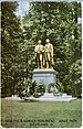 Picture postcard of a large bronze statue of two men on top of a stone pedestal. There are several wreaths that have been placed at the base of the pedestal. The statue is standing in a grassy clearing in front of trees. There is printing along the bottom of the postcard that says "Goethe and Schiller Monument, Wade Park, Cleveland, O."