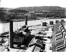 A large dark-coloured rectangular building and a smaller building with three smokestacks. In the background is the river. The steam plant is a small building with two smokestacks.
