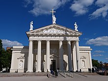 Frontale Farbfotografie einer weißen Kirche mit einem Säuleneingang. An der Fassade sind Figuren und ein langes Relief eingemeißelt. Im Giebeldach ist eine Szene und auf der Dachspitze und an dessen Ecken sind drei Figuren. Die mittlere Figur hält ein goldenes Kreuz.