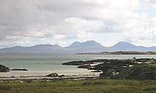 A small sandy beach lies beyond a grassy foreshore. In the distance, the outline of a range of brown and grey hills is visible under cloudy skies.