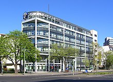 View of a seven-story, modern building, predominantly grey and white, with a cross-like symbol and large letters spelling "Scientology Kirche" at the top.
