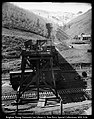 A photograph, taken outdoors, of men working on railroad tracks. There are two sets of tracks running horizontally and one set running up a hill to the distance. There is what appears to be a large chute over the two horizontal tracks. The vertical tracks are likely to be mining tracks with a coal car on them.