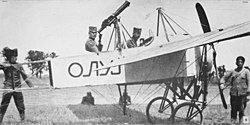 Two men seated in a World War I-era biplane, surrounded by technical personnel.
