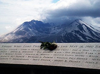 A plaque with the carved names of the eruption's victims appears, with a bouquet of flowers sitting on its center. In the background, Mount St. Helens can be seen.