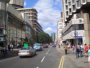 The southern end of Tottenham Court Road in 2005. A blue taxi is in view. There are crowds of pedestrians on either side of the busy street.