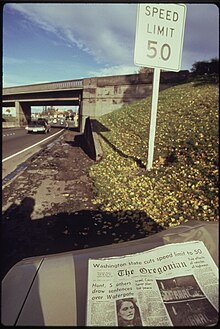 A sign next to a highway says "Speed Limit 50". A newspaper in the foreground has an article about the new speed limit.