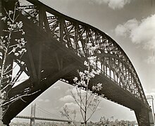 Grayscale image of the main span as seen from immediately beside it, within Astoria Park. Trees in foreground of bridge in bloom, Triborough Bridge and Manhattan visible beyond.