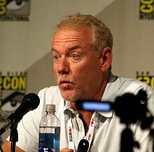 John Debney speaking at ComicCon 2013. He is sitting at a table, with a microphone in front of him and a white backsplash with logos behind him. He is speaking, and in the middle of a hand gesture.