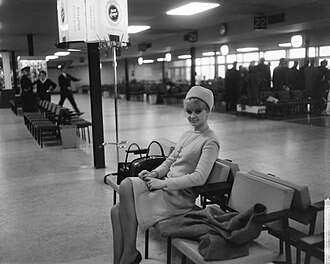 A young woman with short blonde hair turned towards the camera, smiling. She is wearing light clothes and a hat with a crucifix necklace. She is sitting in Amsterdam Airport Schiphol in a waiting chair with her luggage in the seat next to her.