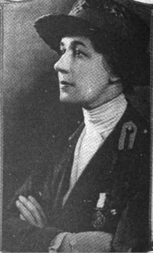 A white woman wearing a hat, arms folded across her chest; she is wearing a high-collared white blouse with a dark jacket; there is a medal pinned to her jacket