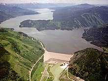 Aerial view looking upstream at an earthen dam and a reservoir stretching off into a mountain valley in the distance