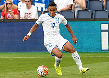 A soccer player wearing a white jersey and white pants dribbling a ball.