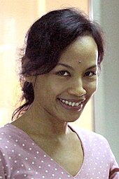An Indonesian woman, looking forward and smiling. She is wearing a purple v-neck shirt