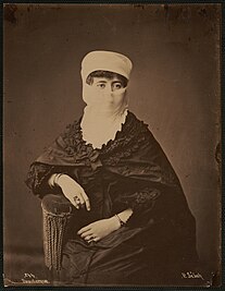 Sepia-toned portrait of a woman seated on a chair, wearing traditional Turkish clothing, including a veil.