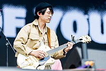 Kim shown in upper body shot, playing his guitar. He wears a light-coloured jacket over a similar coloured shirt and sports a dark cap