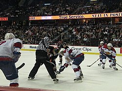 An official wearing black and white stripes conducts a faceoff as several players from two teams anticipate the start of play.
