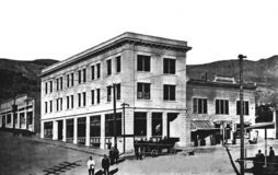 A black and white photograph of the recently opened Cook Bank building in 1908 from the diagonally opposite corner of the intersection. The bank is a three-story white masonry building with many windows on the two visible sides and a door on each side. A cornice wraps around the building at the roofline; the roof is flat. About twelve people are in the street outside the building, along with a car, and three telephone utility poles. A two-story building with awnings is directly attached to the longer side of the bank building (to the right). The road slopes upward to the left, passing a single story building. Hills are visible in the background.