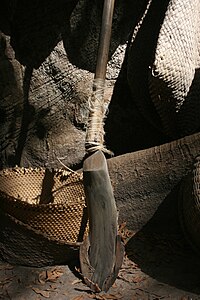A metal blade attached to the base of a wooden rod resting against a tree trunk and various wicker products
