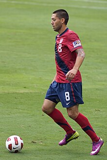 A man wearing a red shirt and blue short pants readies to kick a soccer ball