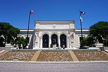 Frontal view of a large grand building. In the foreground a set of fountains that resemble steps are flowing with water. Two flag poles stand on each side of the straiway that leads to the building. There are three large archways with black metal work at the center, and the entrance is in the middle archway. A large outdoor statue blocks the doorway from view. Visitors walk the grounds.