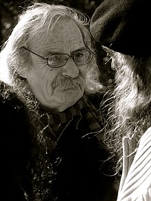 An older looking white man with long hair looking at someone. He is wearing small rectangle glass, and black jacket. You can only see the back of a person who he is looking at. They have long hair, wearing beret and a jacket.