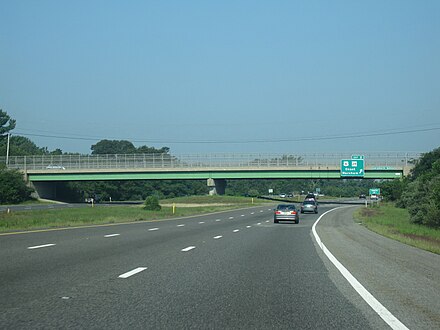 Ground-level view of a three lanes of a divided freeway; a large green and gray overpass bridge and a green exit sign are visible in the distance.