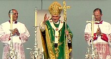 Verschwommene Farbfotografie vom Papst in grün-goldener liturgischer Kleidung mit einem goldenen Kreuzstab in der linken Hand. Er streckt seine rechte Hand zum Segen aus. Neben ihm steht jeweils ein Würdenträger.