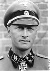 A black-and-white photograph of a man wearing a military uniform, peaked cap and a neck order in shape of an Iron Cross. His cap has an emblem in shape of a human skull and crossed bones.