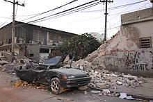 A deep green color car with its roof caved in and the back of the car crushed in. In the background, there is a tree, two phone line poles, and a wall with graffiti on it that is half-broken down.