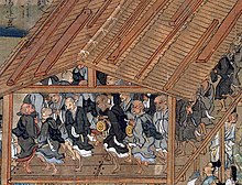In a wooden building open at the sides, a troop of monks in black or grey robes dance in a circle.