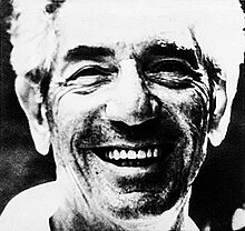 Black-and-white close-up photo of the face of a smiling man with light hair, thick dark eyebrows, and visible stubble.