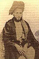 A black-and-white photograph of a man with a dark beard wearing a turban and robes, sitting on a patterned chair, and looking at the viewer