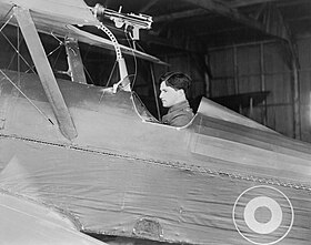 Side view of dark-haired man in open cockpit of biplane equipped with machine-gun on upper wing