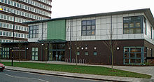 A 2007 two storey library building. The lower floor is brown brick and the upper floor is finished in white square tiles. Above the offset entrance on the left there is a section finished in green. The upper floor has large square windows.