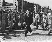 A group of men in suits and military uniforms, marching past, while admiring, a group of saluting soldiers