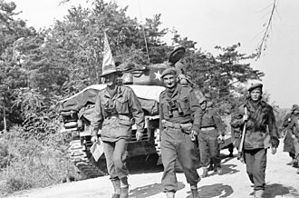 two men in different uniforms walking past a tank, followed by soldiers