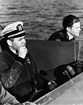Three windswept men at the controls of a boat. One wears a white peaked cap and holds the wheel.