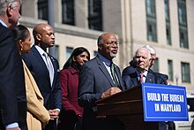 Glenn Ivey speaks at a podium with the text "Build the Bureau in Maryland". He is surrounded by other members of Maryland's congressional delegation, County Executive Angela Alsobrooks, Governor Wes Moore, and Lieutenant Governor Aruna Miller.