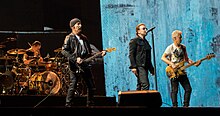 U2 performing on a concert stage. The Edge and Adam Clayton, playing guitars, flank Bono in the foreground, while Larry Mullen, Jr. is behind a drum kit in the background on the left side.