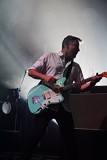 Ben Lloyd, playing a blue electric guitar, in front of a smoky background. Ben is playing on stage, wearing a white open collard shirt. He is lunging while playing the guitar.