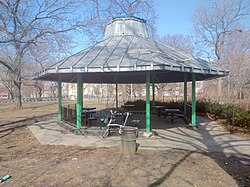 An open-air gazebo. The structure is eight-sided, with a metal roof supported by pillars with decorative iron fencing between the pillars. Under the roof are permanently installed tables with attached seating for about a dozen people.