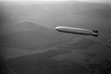 A black-and-white photograph from slightly above of the Graf Zeppelin, a large slim airship, flying from right to left against a low sun. The ground below is misty and consists of rolling hills, woods, and a meandering road which is catching the sun. The silvery airship is also reflecting the sun. It bears black swastikas on white circles on its vertical tail surfaces.