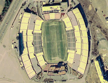Aerial view of Foxboro Stadium with a soccer field surrounded on two sides by three sets of yellow and blue stands, and on two sides by smaller stands.