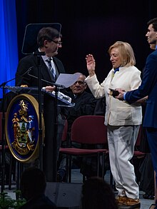 Governor Janet Mills in a white pants suit and LL Bean boots with her left hand on a bible and her right hand raised. She is surrounded by other state government employees on a stage.