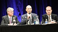 From left to right, a bespectacled middle aged man, a younger bald man holding a wireless microphone, and a balding middle aged man, all in suits, sit at a long dark table in front of dark blue curtains.