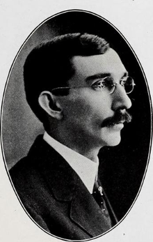 A black-and-white headshot photo of a bespectacled, mustached man wearing a suit and tie