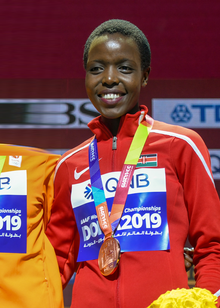 Agnes Tirop wearing a red athletic Nike jacket with a Kenyan flag, a 2019 World Athletics Championships placard, as well as her bronze medal, standing on a podium next to another contestant (not seen in photo), holding a bouquet of yellow flowers, grinning at audience.