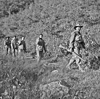 Three unarmed soldiers being led down a hill by two soldiers armed with rifles, one in the front the other in the rear.