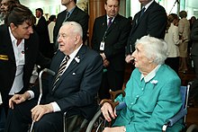 Whitlam, in extreme old age, sits with an elderly lady as a woman bends to speak with him. He holds a metal cane. Other people, mostly men, stand behind him.